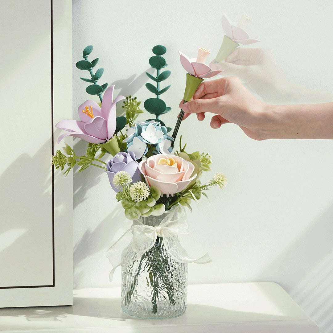 Hand arranging artificial flowers in a clear vase on a white surface.