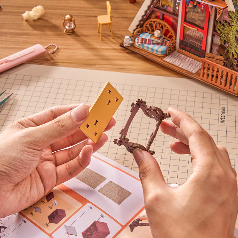 Hands holding a miniature yellow power outlet model and a brown wire frame piece over an instruction sheet on a wooden table, with a tiny house and lantern in the background.