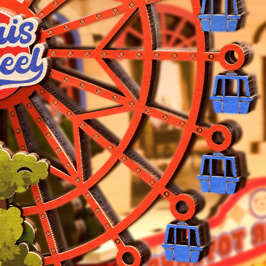 A close-up of a colorful Ferris wheel cutout or decorative sign, showing red-orange wheel structure, blue gondolas, and part of a sign that reads "Ferris" against a blurred warm-toned background.