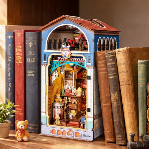 A miniature toy bookshop diorama model on a wooden shelf, flanked by vintage hardcover books, with a small teddy bear and potted plant in the foreground.