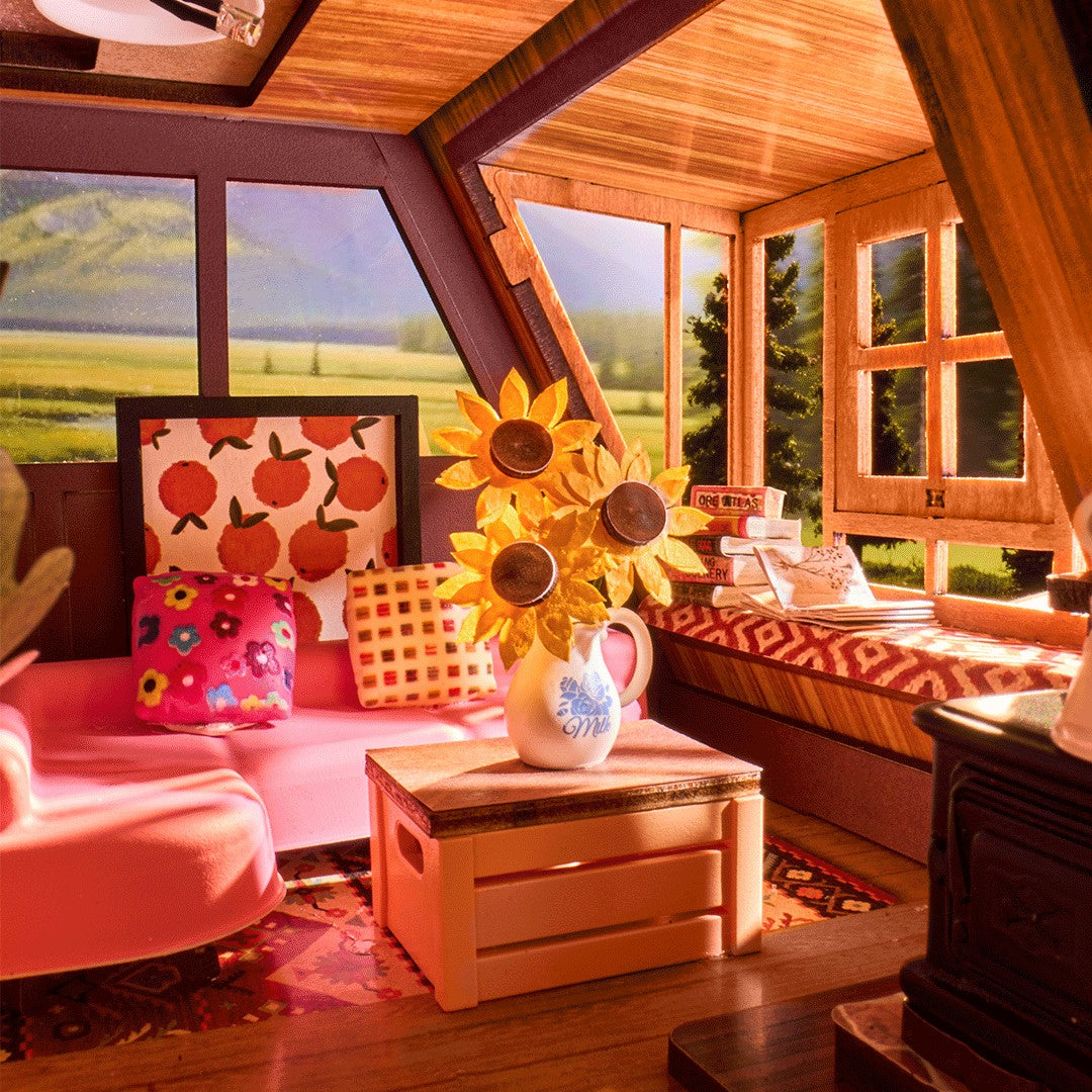 A cozy miniature dollhouse interior with pink seating, patterned pillows, a wooden crate table, a white pitcher filled with yellow sunflower decorations, and large windows showing a painted outdoor landscape.