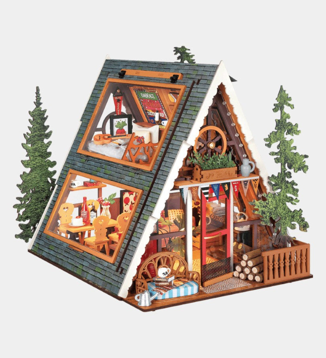 A detailed miniature A-frame cabin dollhouse with a gray shingled roof, warm wood-toned walls, and tiny furnished rooms visible through large windows, flanked by small green pine trees on a light gray background.