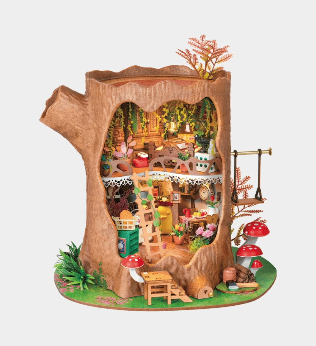 A miniature fairy-tale treehouse dollhouse set inside a hollow tree trunk, featuring tiny furniture pieces, a small kitchen, shelves, hanging plants, and red-and-white mushrooms on a green base, shown against a plain light gray background.