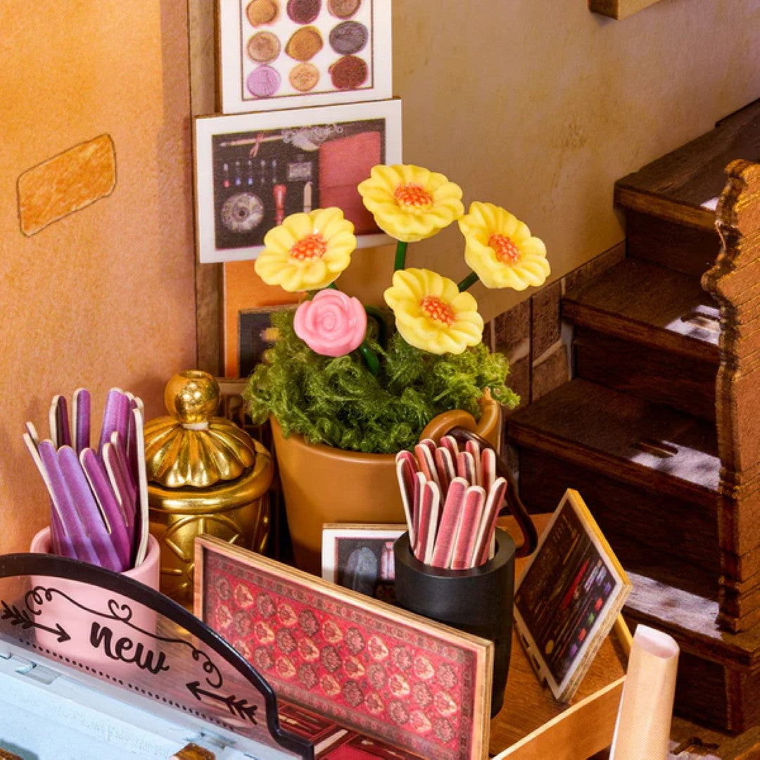 A miniature dollhouse-style room scene with a potted flower arrangement in the center, yellow and pink artificial flowers in a terracotta pot, small decorative frames, a gold lantern, and miniature furniture against a warm tan wall.