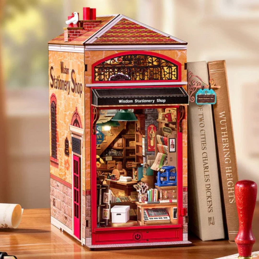 A miniature house-shaped model of a vintage stationery shop sits on a wooden table, styled with books, a fountain pen, and a red wax stamp. The shop features a detailed interior visible through a large window, with red trim and a gabled roof.