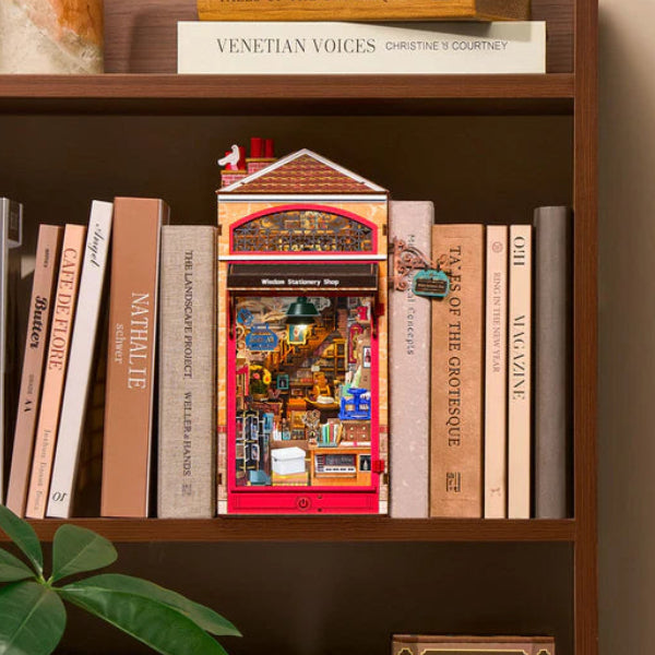 A miniature red and gold Victorian-style house bookshelf decoration displayed on a dark brown wooden bookshelf next to books, with a green plant leaf in the foreground.